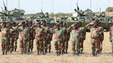A group of Zimbabwean soldiers stand in salute in a grassy field. They are wearing camouflaged uniform and black berets. Behind them, six tanks are positioned. Its hulls are painted a dark green. Further back, a line of trees are seen over the horizon. A light blue sky serves as the background.