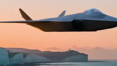 A grey stealth fighter jet is seen flying in front of a shoreline during sunset. A lighthouse close to the white cliff face is seen over the horizon. The sky is a warm orange color.