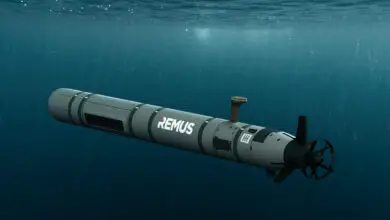 A computer-generated model of a REMUS medium-class unmanned underwater vehicle (UUV) is seen submerged in blue-green waters. It has a cylindrical shape, with propellers on its back end. The greenish-grey hull has the white square HII monogram painted on it, as well as the word 'REMUS'.