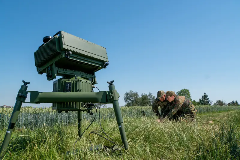 A Thales surveillance system is seen propped up in a grassy plains. It's positioned on the left side of the foreground, with two military personnel in camouflage on the far right in the background. The sky is a clear, cloudless blue.