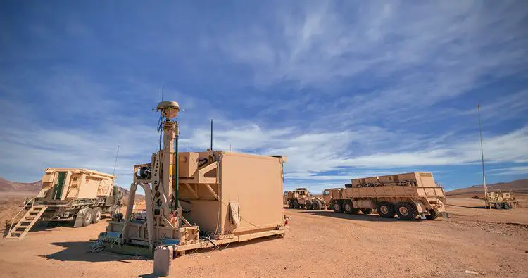 An Engagement Operations Center (EOC) is seen at a desert landscape at White Sands Missile Range, New Mexico. The center is composed of three trucks and one command station. The center's paint shares the same red-orange hue as the desert landscape. The background is a clear blue sky, dotted with a few thin clouds.