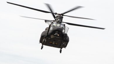 A black MH-47G Block II undergoing flight test is seen mid-flight. Its two overhead rotary blades are a little blurred as they spin. The background is a clear white sky.