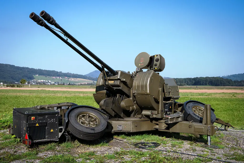 An Oerlikon GDF 103 anti-aircraft gun is seen set up in a grassy plains. Its two gun barrels are pointed upwards to the left at a 45-degree angle. The whole gun system is painted a deep green. The far background is a clear, blue sky.