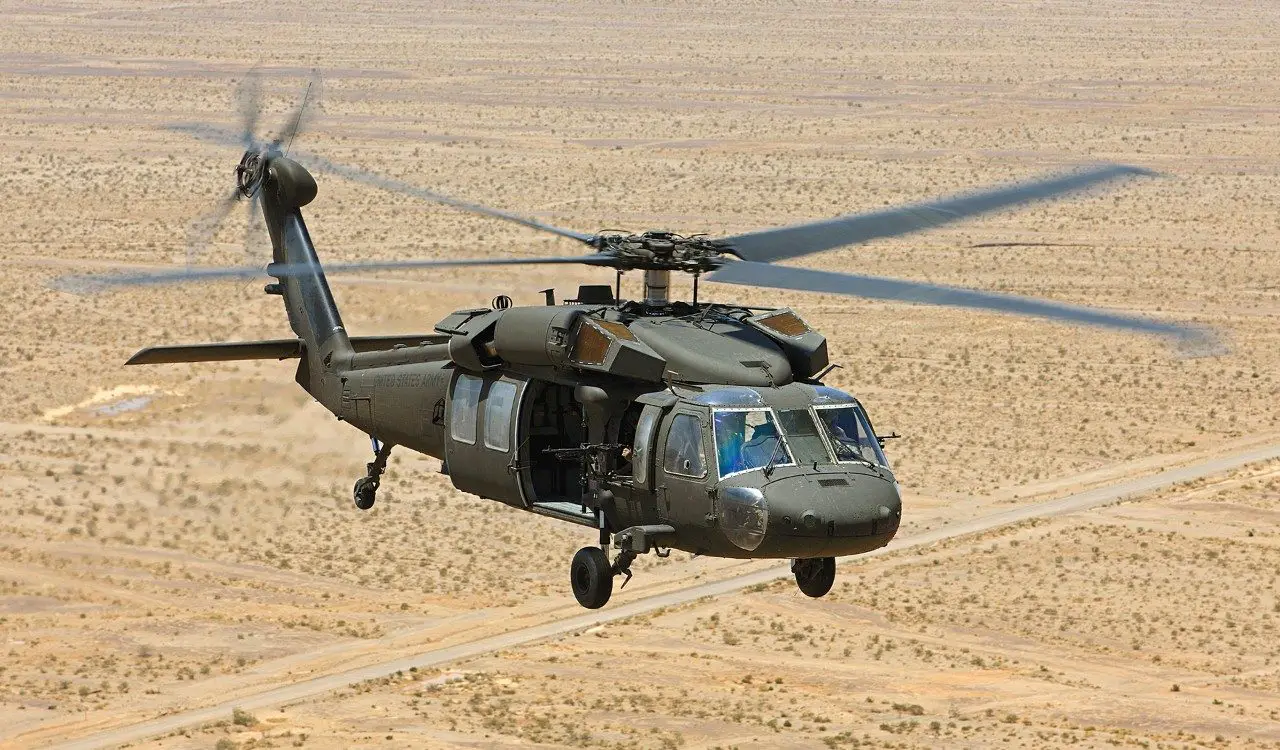 A Sikorsky UH-60M Black Hawk helicopter is seen flying over a desert plains. The dry, almost barren ground below has a pale yellow color. The helicopter's four spinning blades are blurry from its rotation. The helicopter's paint is dark green.