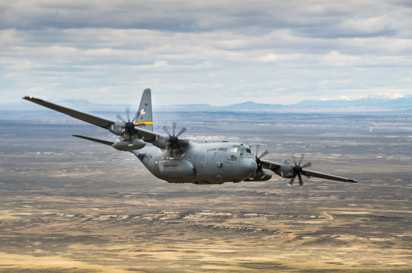 A C-130 Hercules flies over a sandy plains. The plane is colored gray, with a yellow stripe on its tail. It has "US AIR FORCE" painted on its side in black, as well as "WYOMING AIR GUARD", below its wing. The background is the landscape; a wide stretch of land occupies most of the space, with green grass and yellow-white sands scattered evenly. Over the horizon, a mountain range is seen. Some of its peaks are snowy and white. The sky above is full of thin, long clouds.