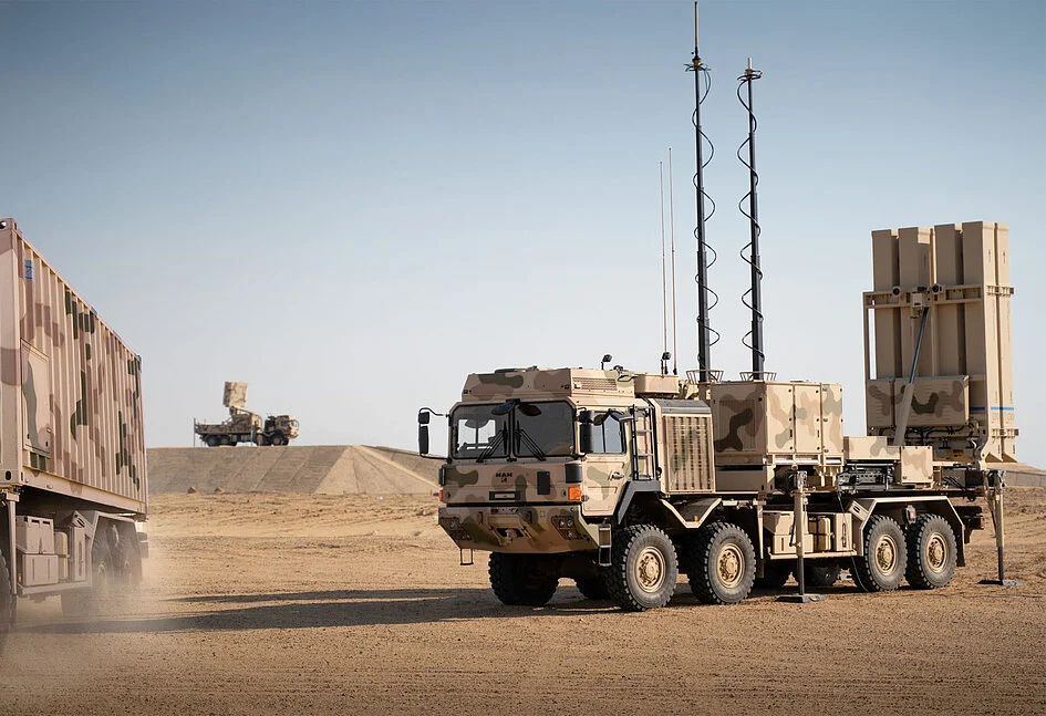 An IRIS-T SLM air defense system is seen in what appears to be a dry/desert landscape. Some sand can be seen being blown by the wind. The self-launching system itself is seen propped up and mounted on the back of an 8x8 military truck. The truck is painted in camouflage that matches the dry brown color of the environment. The background is a clear but dull blue sky.