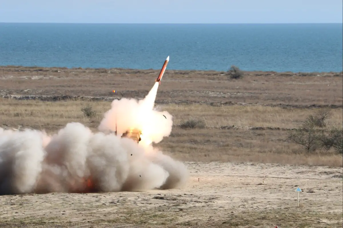 A missile from the Patriot missile system takes off in the middle of a field. The red missile is seen beginning to blast off, leaving a trail of smoke behind and a jet of white-orange flames. The background is a calm and blue body of water that stretches to the horizon, and a light-blue, cloudless sky.