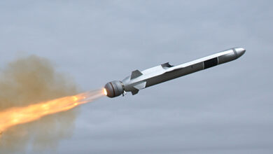 A Kongsberg Naval Strike Missile is seen propelling itself in the air. Its long body is painted metallic silver, and hot orange flames are seen jutting out of its back. A faint orange-grey smoke is seen around its flames. The background is a dull blue sky.