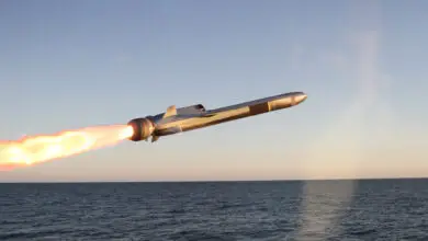 Kongsberg's Naval Strike Missile (NSM) is seen front and center, launching from the left. A jet of red-orange flame streaks from its back. The background is a deep blue ocean, with a cloudless sky farther back.