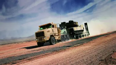 An Oshkosh Heavy Equipment Transporter (HET) A1 truck is seen moving across a sandy area. It leaves behind a dusty trail as it moves to the left. It's painted dirty brown, and on its back is an armored tank painted in brown and gray camouflage. The sky is a deep blue with streaks of white clouds.