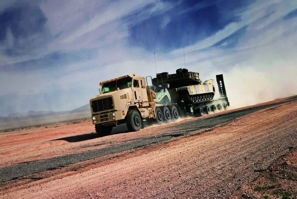 An Oshkosh Heavy Equipment Transporter (HET) A1 truck is seen moving across a sandy area. It leaves behind a dusty trail as it moves to the left. It's painted dirty brown, and on its back is an armored tank painted in brown and gray camouflage. The sky is a deep blue with streaks of white clouds.