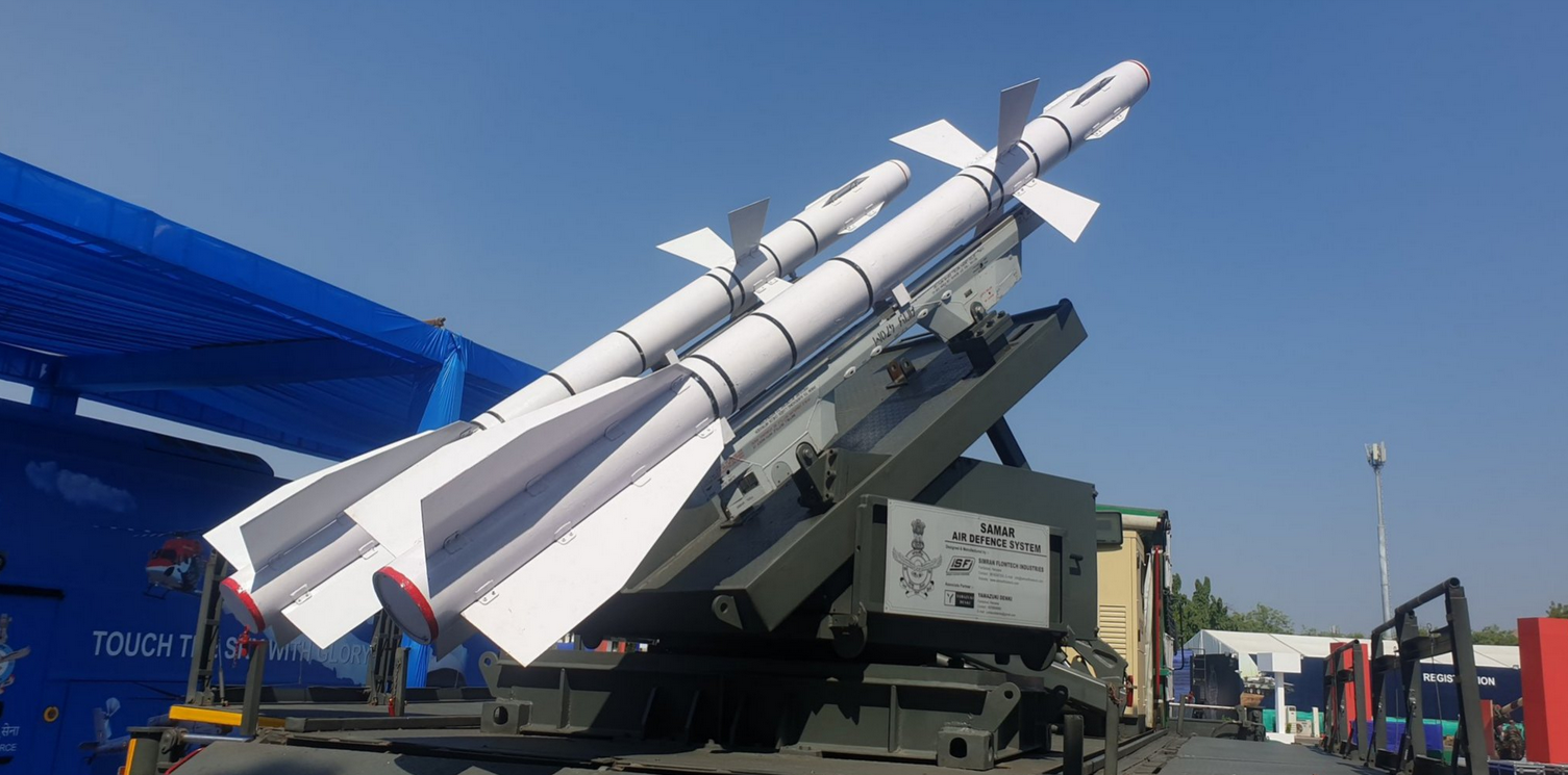The Surface-to-Air Missile for Assured Retaliation (SAMAR) missile ssytem is seen in the middle of the frame. The launcher is painted a deep green. Two missiles, painted white, are propped on it, pointing upwards. The background is a light blue sky.