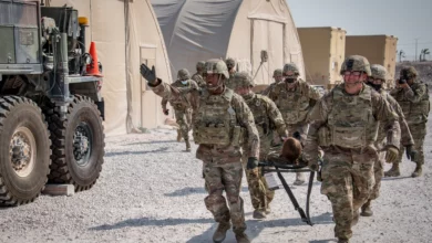 Four camouflaged US Soldiers at the Al Udeid Air Base in Qatar are seen carrying a soldier on a stretcher in what appears to be a military drill. On the back, at least six more soldiers are seen tailing them. On the background is two brown tents and two brown military trucks. On the left, the back of another truck is seen.