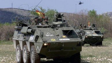 Two dark-green BMR-600 armored vehicles are seen in a grassy plains. The one in the foreground has three camouflaged soldiers on it, while the one on the background has two. Both vehicles have the Spanish flag propped up on its back, and have what appears to be protective barbed wire wrapped around them. In the far background, a helicopter can be seen flying on the dark-blue sky.