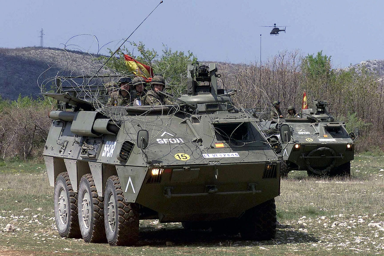 Two dark-green BMR-600 armored vehicles are seen in a grassy plains. The one in the foreground has three camouflaged soldiers on it, while the one on the background has two. Both vehicles have the Spanish flag propped up on its back, and have what appears to be protective barbed wire wrapped around them. In the far background, a helicopter can be seen flying on the dark-blue sky.