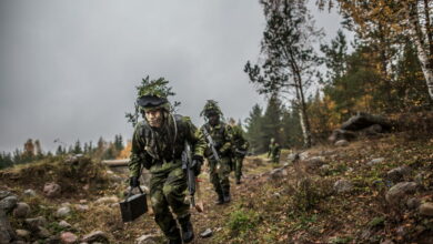 Four Swedish Armed Forces soldiers walk while crouching in a rocky forested area. They are seen wearing camouflage with leaves on top of their helmets to blend in more with the green and gloomy environment. All of them are armed with rifles. The background is a sky fully covered by a deep gray cloud.