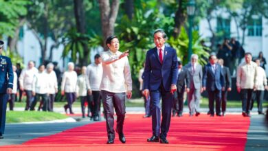 Philippine President Ferdinand Marcos Jr. and Indonesian President Joko Widodo are seen side-by-side as they walk on a red carpet in the grounds of Malacañang Palace in Manila, Philippines. Several men in military uniform, suits, and barong are seen walking behind them. A few trees are seen in the background.