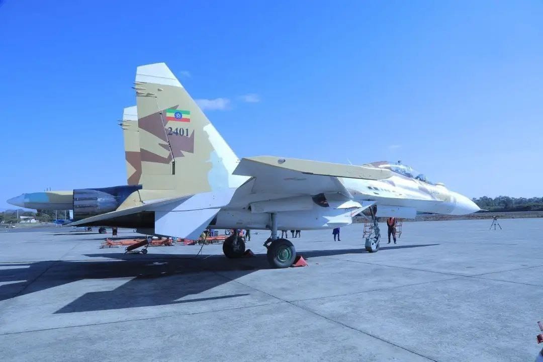 Two Sukhoi Su-30 planes are seen stationed on what appears to be a military air base. The planes are painted white, except for its camouflaged tails. The plane in front has "2401" painted on its tail, as well as the flag of Ethiopia. The background is a clear blue sky.