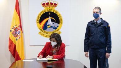 Spanish Defense Minister Margarita Robles is seen signing a document inside what appears to be a conference room. An office personnel is seen standing by her left. The Spanish Air and Space Force emblem is displayed on the background, and the Spanish flag is positioned to her right.