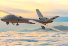 A gray General Atomics MQ-9B SkyGuardian is seen flying above the clouds. On the far right side of the background, a mountain range is seen peeking out. The scenery is tinged in the setting sun's orange hues.