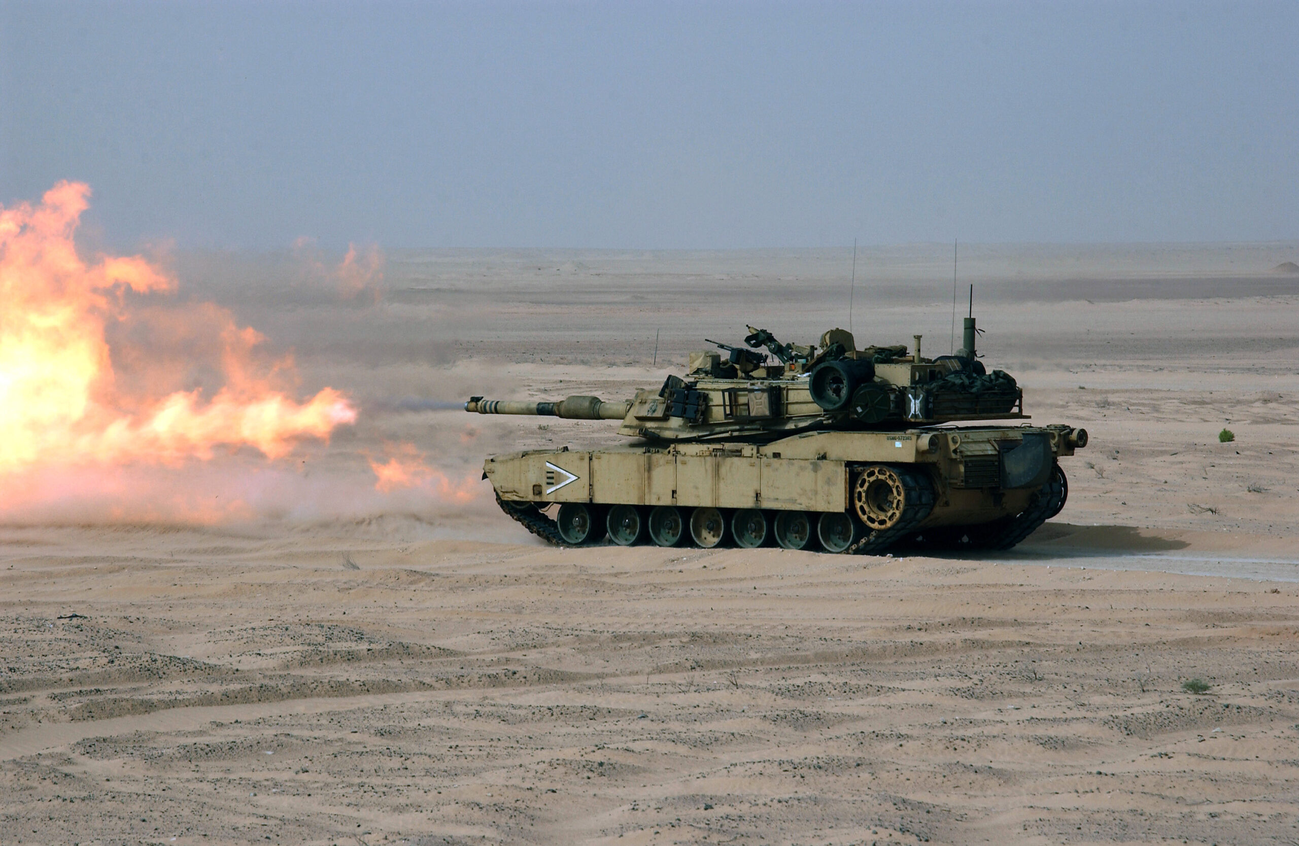 An Abrams M1A1 tank is seen initiating live fire in a desert field. Red-orange flames appear to be coming out of its turret. The tank's hull blends well with the area's dry, sandy environment. The background is a dim blue sky.