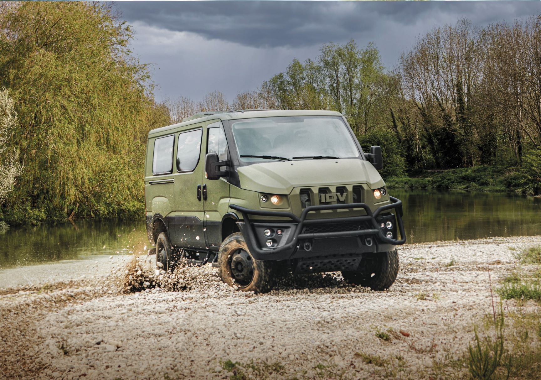 An Iveco Military Utility Vehicle is seen traversing a flooded and muddy terrain. The camouflage-green van's wheels are covered in mud, and the brown water on the ground is seen rippling and splashing because of the vehicle. The area appears to be a swamp or a forest, with overgrown bushes and trees in the background. The sky is dark and cloudy.