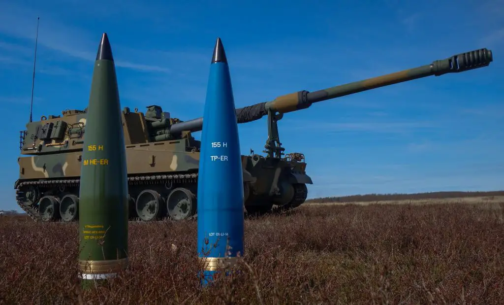 Two 155mm artillery ammunition are placed upright in a grassy field. The one on the left has its shell painted green, and the other is paited blue. A camouflage-painted battle tank is stationed behind them. The background is a clear blue sky.