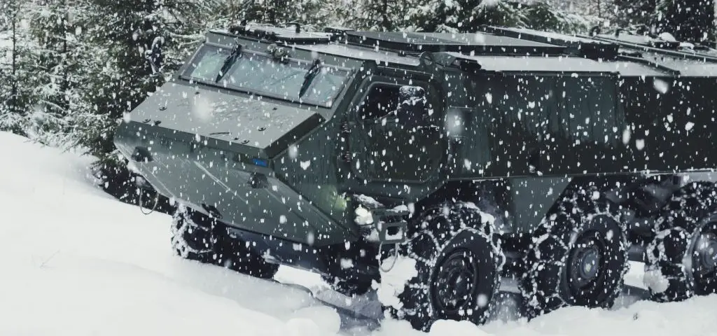 A dark green Patria 6x6 armored vehicle is seen traversing a snowy field while it's snowing. Trees can be seen on the background, covered in white snow. The vehicle's wheels are fitted with snow chains to help it have better grip and control on the ground.