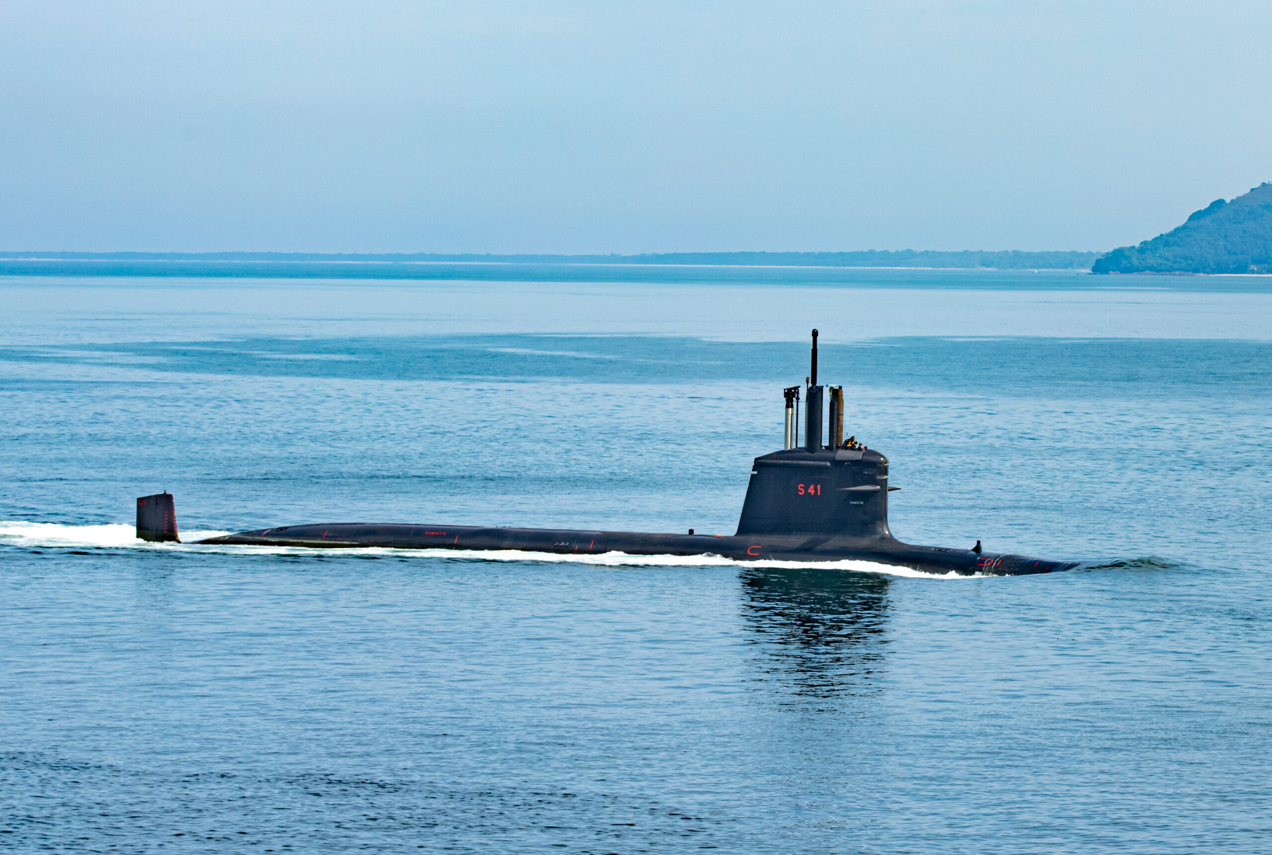 The black Humaitá (S-41) submarine is seen breaking the surface of the sea. Near the top of its hatch, "S-41" is painted in red. The sea and the sky are a calm blue.