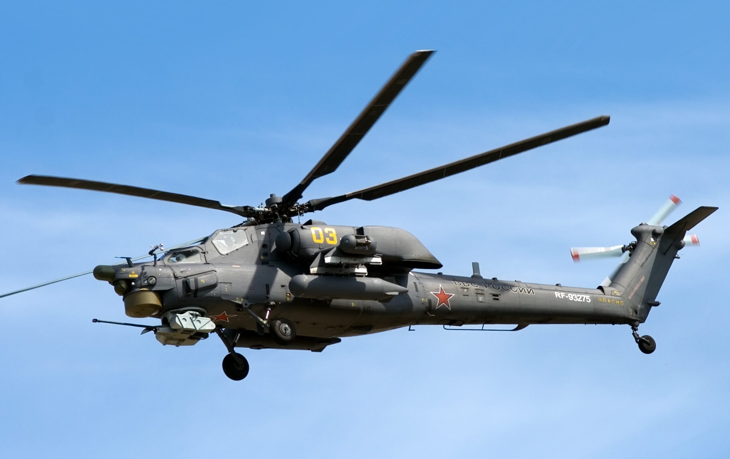 A Mil Mi-28 helicopter is seen hovering on the clear blue sky. On its gray-green hull, the number "03" is painted in yellow, as well as two red stars and the unit name "RF-93275" in white. Some characters on the Russian alphabet are also painted in black.