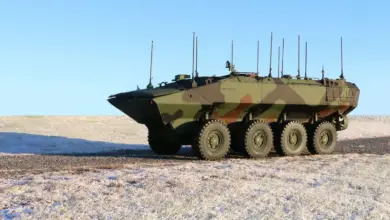 A camouflage-painted Amphibious Vehicle Command and Control (ACV-C) variant is seen stationed in a dry plains area. The eight-wheeled vehicle has 12 antennas installed on it, all pointing upwards. The background is a solid baby-blue sky.
