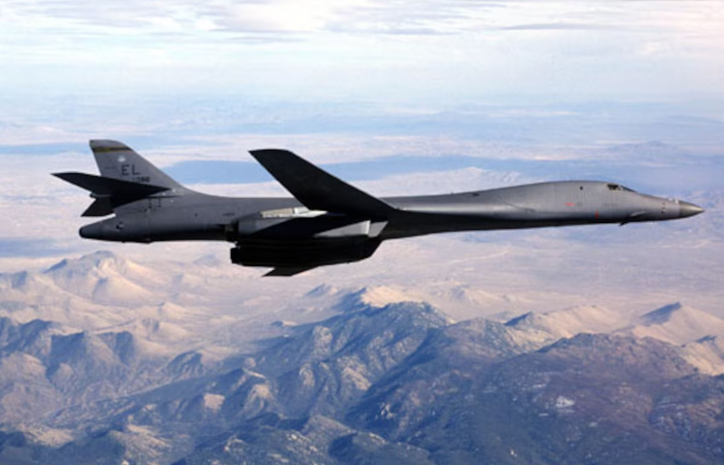 A black B-1B Lancer bomber is seen flying above a mountainous region. The mountains' land have colors ranging from forest's greens to desert yellows and whites. The sky above is cloudy and white.