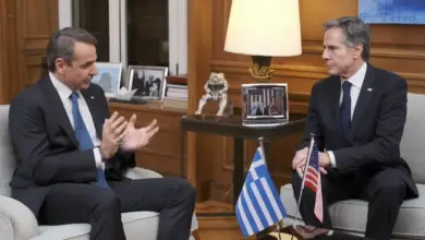 Greek Prime Minister Kyriakos Mitsotakis is seen speaking with US Secretary of State Anthony Blinken. They' re sat across each other, with a table in the middle decorated with the Greek and US flags.