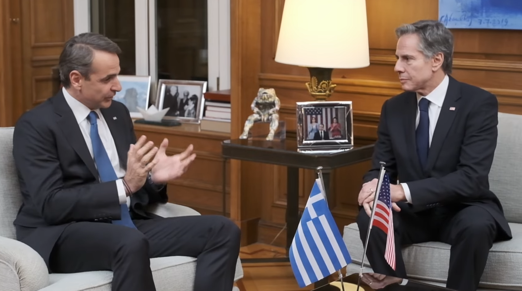 Greek Prime Minister Kyriakos Mitsotakis is seen speaking with US Secretary of State Anthony Blinken. They' re sat across each other, with a table in the middle decorated with the Greek and US flags.