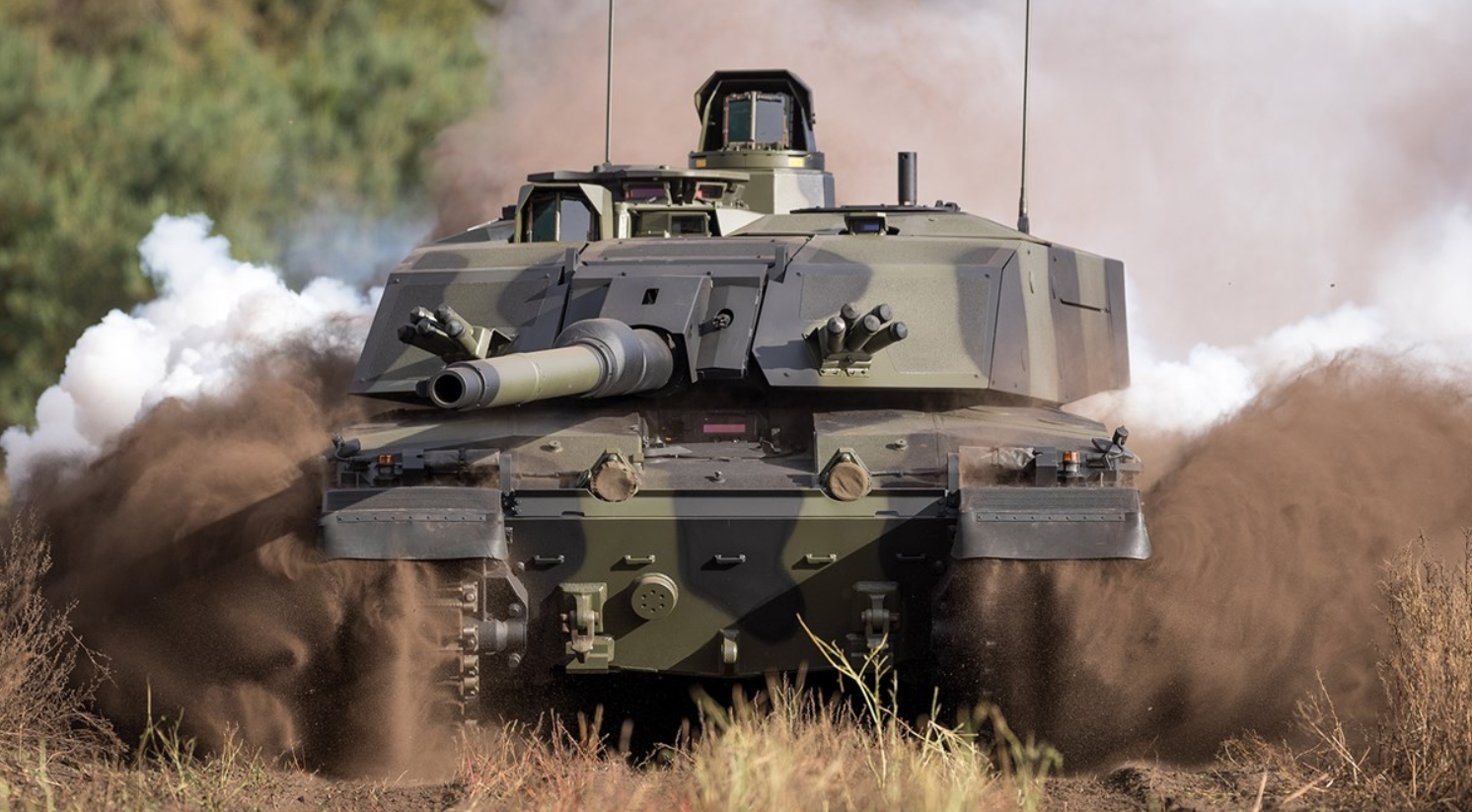 The British Army's Challenger 3 Main Battle Tank is seen moving across a dusty grassland. The tank is painted in black and green camouflage, and its tracked wheels are shrouded in brown dust from the ground. In the back, white smoke is seen, presumably from the tank's exhaust.