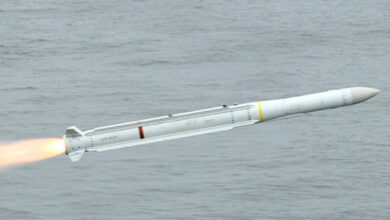 A Raytheon Sea Sparrow missile is seen flying over gray waters. The white missile is propelled by its engine, jutting out white-to-red-hot flames on its back.