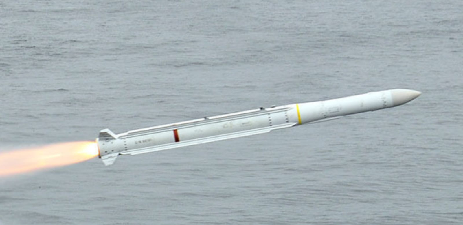 A Raytheon Sea Sparrow missile is seen flying over gray waters. The white missile is propelled by its engine, jutting out white-to-red-hot flames on its back.