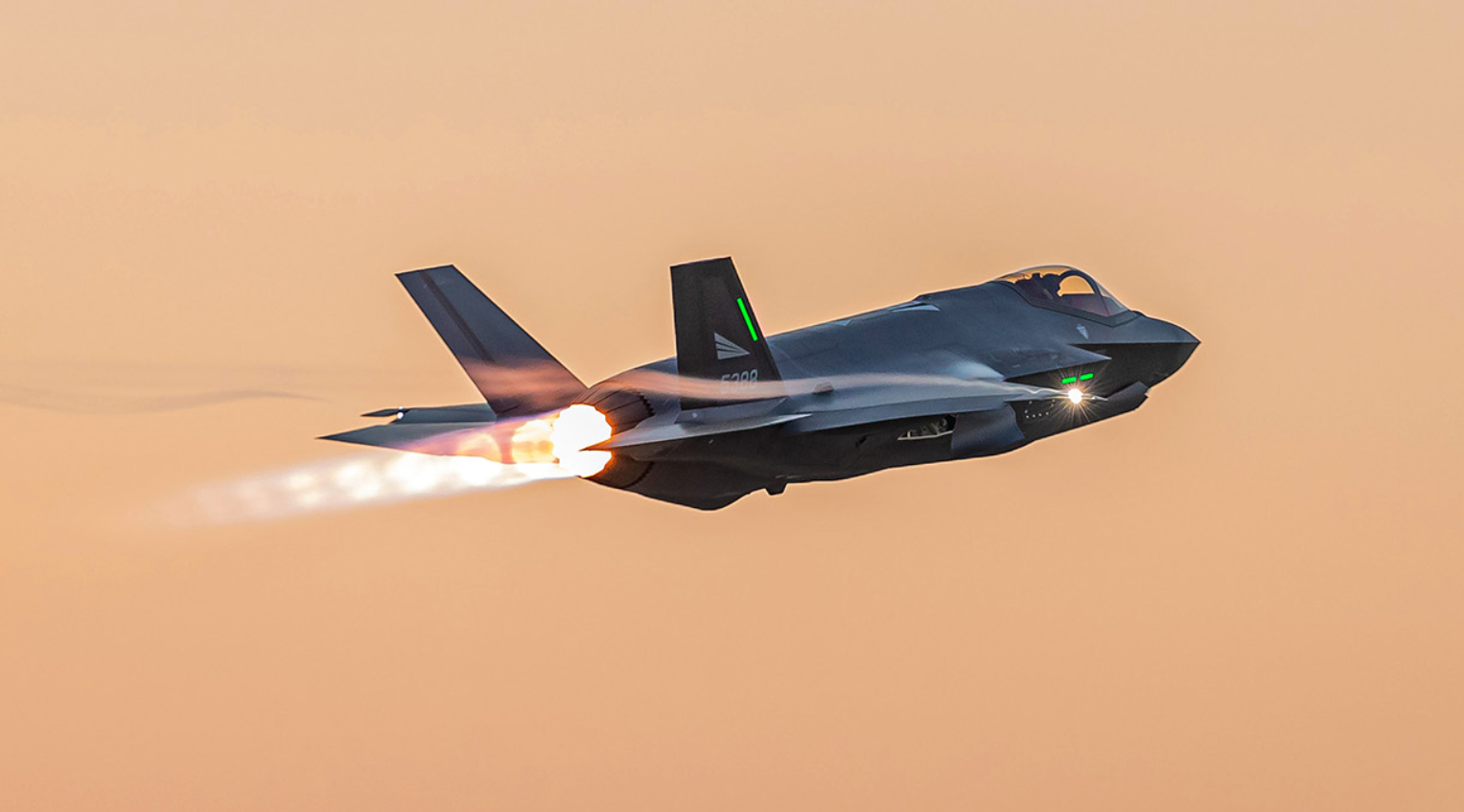 A black Royal Norwegian Air Force F-35 plane is seen flying in an orange, sunset-tinged sky. The plane's wing has "5388" painted on it in white. A trail of red-hot jet flames is seen coming out of its back.