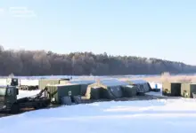Hospitainer's Role II Hospital, a mobile, inflatable building, is seen set up in a snowy field outside a forested area in the background. Two container trucks are parked outside of it, and two cargo truck containers are also positioned on the far right of the field.