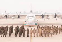 Uniformed soldiers from the Indian and Saudi Arabian Army stand in attention in front of a white plane in what appears to be an airport runway. Between the soldiers and the aircraft, a huge banner reads 'EX SADA TANSEEQ - 2024'. India's flag is on the leftmost side of the banner, while Saudi Arabia's is on the right. The background is a foggy gray sky with a barely visible skyline in the distance.
