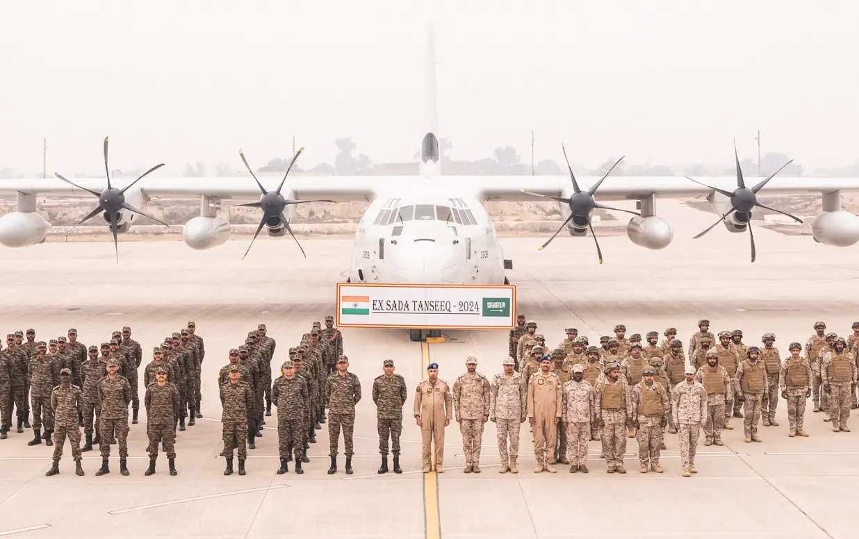 Uniformed soldiers from the Indian and Saudi Arabian Army stand in attention in front of a white plane in what appears to be an airport runway. Between the soldiers and the aircraft, a huge banner reads 'EX SADA TANSEEQ - 2024'. India's flag is on the leftmost side of the banner, while Saudi Arabia's is on the right. The background is a foggy gray sky with a barely visible skyline in the distance.