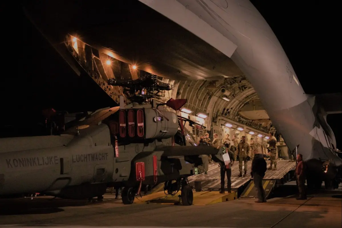 A gray AH-64E Version 6 Apache attack helicopter is seen being unloaded out from a US Air Force C-17 airlifter via a ramp. Ten individuals, seven of whom are wearing their camouflaged uniforms, are on the scene, occupied with the delivery of the aircraft.