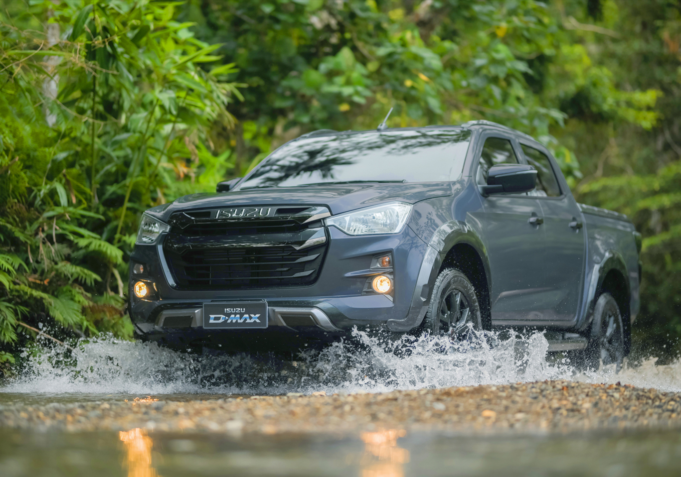 A gray Isuzu D-Max is seen being driven across a shallow pool of water in what appears to be an off-road location, with uneven ground and a background of green-leaved trees. The clear-white water around the truck's wheels are seen splashing towards all directions.