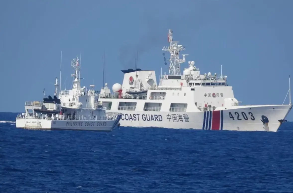 A white Philippine Coast Guard ship is stationed in the waters of the West Philippine Sea, facing a larger Chinese Coast Guard ship. The sea is a deep blue color, and the sky is a dull blue.