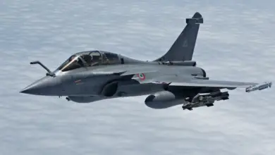 A gray Dassault Rafale fighter jet is seen flying above a sea of white clouds. It's equipped with what appears to be two missiles under the middle of its left wing, and a smaller one on its tip.