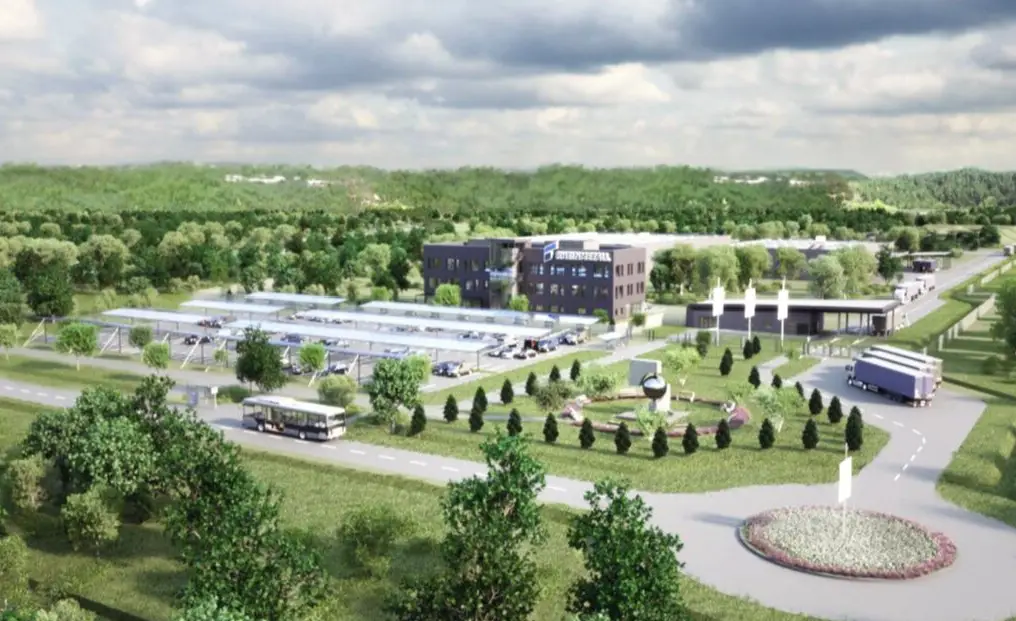 An artist's rendering of Rheinmetall's Várpalota plant. The complex shows a roofed parking lot three buildings, and what appears to be a small park. Three trucks and a bus are seen parked by the perimeter of the site. A roundabout is also shown on the lower right corner of the image. Most of the surrounding area is dotted by trees and grass.
