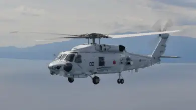 A Mitsubishi SH-60L is seen flying above the sea. The white-gray helicopter has "02" painted in black on its side. The background shows a mountainous shoreline, as well as the calm waters in its front. The sky is slightly gloomy and cloudy.