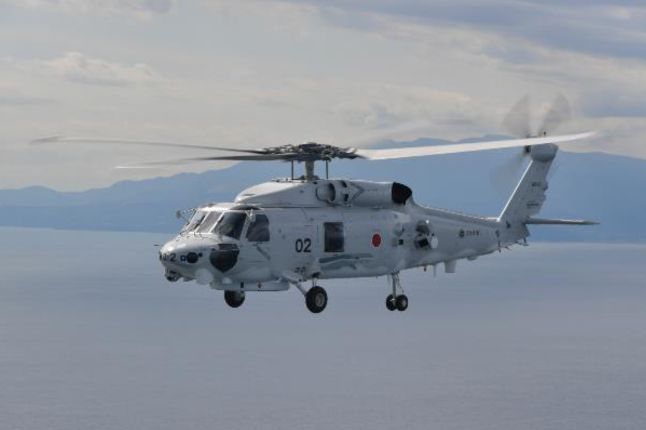 A Mitsubishi SH-60L is seen flying above the sea. The white-gray helicopter has "02" painted in black on its side. The background shows a mountainous shoreline, as well as the calm waters in its front. The sky is slightly gloomy and cloudy.