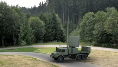 A Thales Ground Master 200 Multi-Mission Compact radar is seen parked in a field with trees in the back. The green-colored truck has a tall antenna propped on its back, with a panel that appears to be the radar.