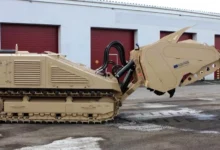 A DOK-ING MV-10 mine-clearing vehicle can be seen outside of a warehouse. The brown-painted armored vehicle has tracked wheels and a tiller mechanism on its front. On the right, a camouflaged soldier is seen standing and looking at it.